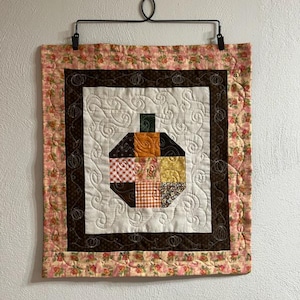 May include: A quilted wall hanging featuring a patchwork pumpkin design. The pumpkin is composed of various patterned fabrics in orange, yellow, and brown. The quilt has a floral border and is suspended from a black metal hanger.