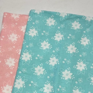 May include: Two fabric pieces, one pink and one teal, are displayed. Both fabrics feature a white floral pattern with detailed flower and leaf outlines. The floral design is repeated across the fabric, creating a consistent pattern.