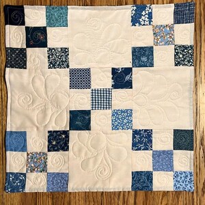 May include: A square quilt featuring a patchwork design of cream-colored fabric blocks alternating with various blue patterned squares. The cream blocks are decorated with intricate, stitched floral and swirling patterns throughout.