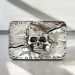 May include: A silver belt buckle with a skull design. The skull is cracked and has a textured surface. The buckle is surrounded by a row of small circles.