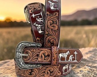 May include: A dark brown leather belt featuring intricate floral tooling and a silver-toned metal buckle. The belt displays a repeating white graphic of a person kneeling beside a saddled horse and a cross, with white stitching along the edges.