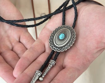 May include: A silver bolo tie with a turquoise stone in the center of a decorative oval pendant. The tie is made of black braided leather.