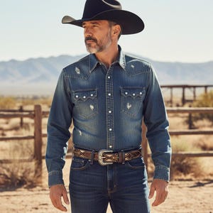 May include: A denim shirt with distressed details, paired with dark wash jeans and a decorative belt with a silver buckle. The outfit is completed with a black cowboy hat. The scene is set outdoors with a desert landscape in the background.
