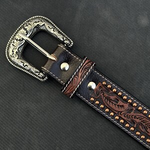May include: Close-up of a brown leather belt with a silver-colored buckle. The belt features a detailed floral pattern and small, gold-colored studs. The buckle has an ornate, engraved design.