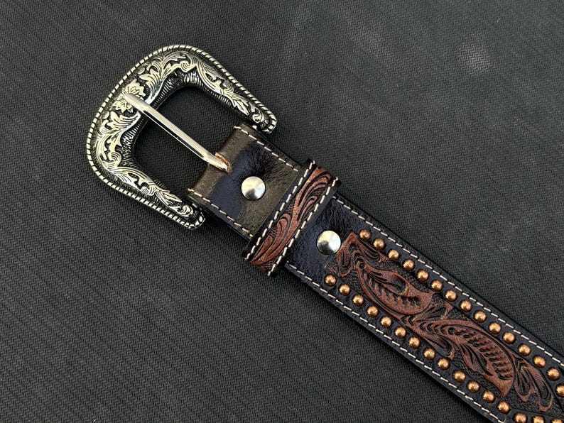 May include: Close-up of a brown leather belt with a silver-colored buckle. The belt features a detailed floral pattern and small, gold-colored studs. The buckle has an ornate, engraved design.