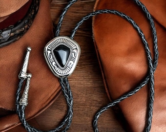 May include: A silver bolo tie with a black stone and intricate design. The tie is made of braided leather and has a silver clasp.