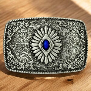 May include: Silver belt buckle with an ornate design featuring a blue gemstone in the center.