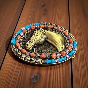 May include: An oval belt buckle with a horse head design. The buckle has a gold-coloured horse head in the centre, framed by turquoise and orange beads. The metal buckle has a classic Western style, suitable for a belt. The buckle measures approximately 8 cm.