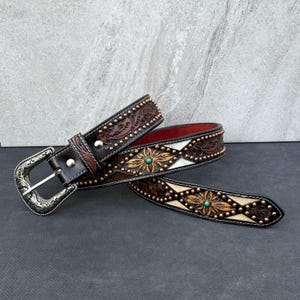 May include: A brown leather belt with floral and geometric patterns, turquoise accents, and a silver buckle. The belt features a red interior and is adorned with small metal studs. The design is reminiscent of Western wear.