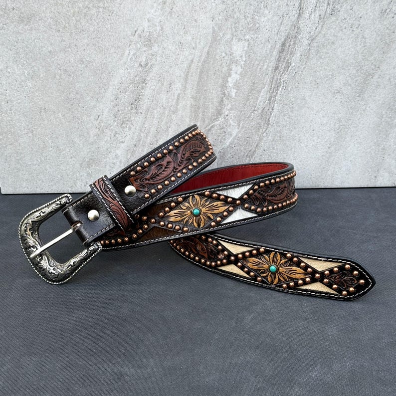 May include: A brown leather belt with floral and geometric patterns, turquoise accents, and a silver buckle. The belt features a red interior and is adorned with small metal studs. The design is reminiscent of Western wear.