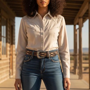 May include: A beige long-sleeve button-down shirt with embroidered floral details and a decorative belt with a silver buckle. The shirt is paired with blue jeans. The belt is brown with a decorative pattern.
