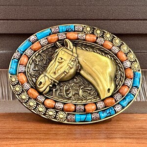 May include: An oval, gold-coloured belt buckle with a horse head design. The buckle is decorated with turquoise and orange stones around the edge. The horse head has a bridle and is set against a decorative background.