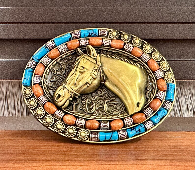 May include: An oval, gold-coloured belt buckle with a horse head design. The buckle is decorated with turquoise and orange stones around the edge. The horse head has a bridle and is set against a decorative background.