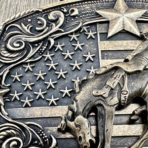 May include: A bronze-toned belt buckle featuring a cowboy riding a bucking bronco against a backdrop of the American flag. The buckle has a detailed design with stars, stripes, and decorative scrollwork, evoking a Western theme.