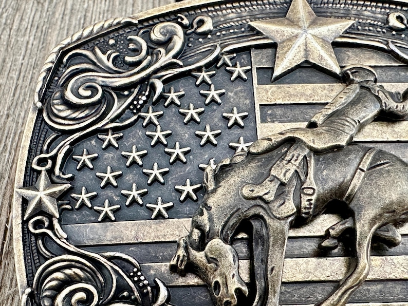 May include: A bronze-toned belt buckle featuring a cowboy riding a bucking bronco against a backdrop of the American flag. The buckle has a detailed design with stars, stripes, and decorative scrollwork, evoking a Western theme.