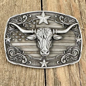 May include: Silver-toned metal belt buckle with a longhorn steer design. The buckle features an American flag motif with stars and stripes, and decorative scrollwork around the edges. The longhorn's head is centered, with large horns extending outwards.