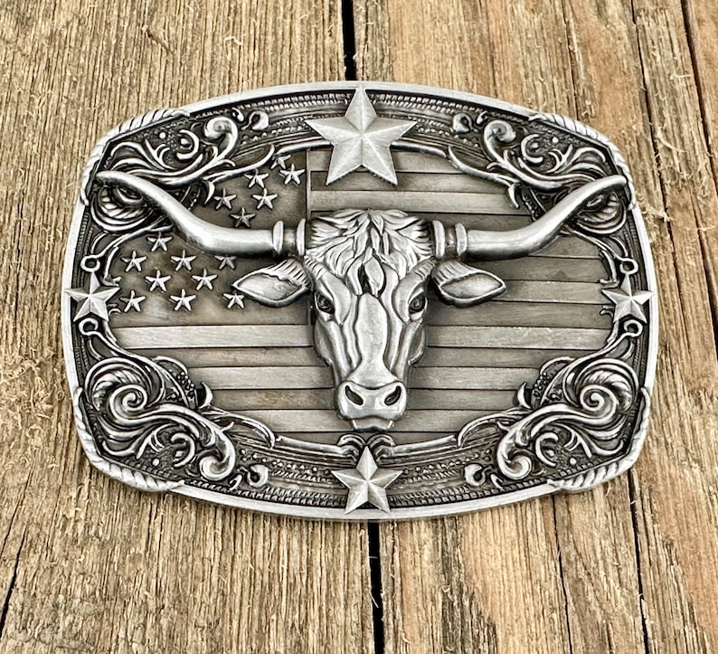 May include: Silver-toned metal belt buckle with a longhorn steer design. The buckle features an American flag motif with stars and stripes, and decorative scrollwork around the edges. The longhorn's head is centered, with large horns extending outwards.