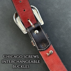 May include: Close-up of a leather belt with a red interior and black exterior. The belt features a silver buckle and Chicago screws for interchangeable buckles. The text "CHICAGO SCREWS INTERCHANGEABLE BUCKLES" is visible.