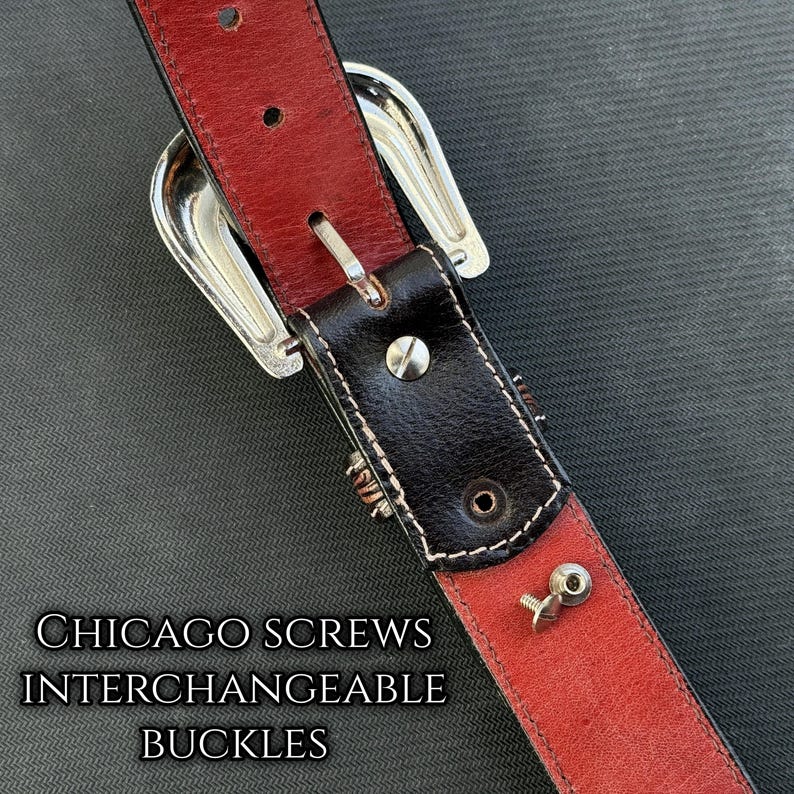 May include: Close-up of a leather belt with a red interior and black exterior. The belt features a silver buckle and Chicago screws for interchangeable buckles. The text "CHICAGO SCREWS INTERCHANGEABLE BUCKLES" is visible.