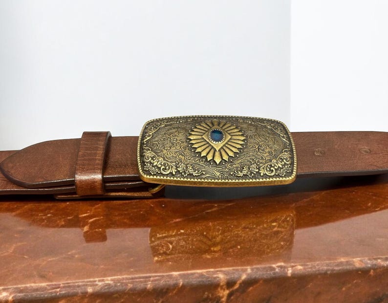 May include: A brown leather belt with a gold-toned buckle featuring an ornate design and a blue gemstone in the center.