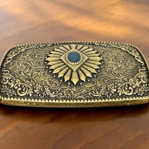 May include: A gold-toned belt buckle with an ornate design featuring a blue gemstone in the center.