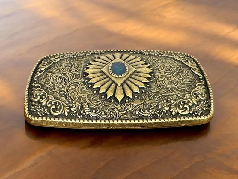 May include: A gold-toned belt buckle with an ornate design featuring a blue gemstone in the center.
