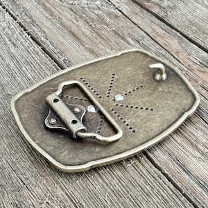 May include: A rectangular, antique brass-colored belt buckle. The buckle features a prong and a loop for the belt. The back of the buckle has a series of small holes and two clear, round embellishments. The buckle is set against a weathered wooden surface.