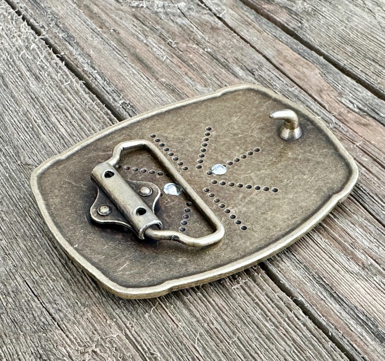 May include: A rectangular, antique brass-colored belt buckle. The buckle features a prong and a loop for the belt. The back of the buckle has a series of small holes and two clear, round embellishments. The buckle is set against a weathered wooden surface.