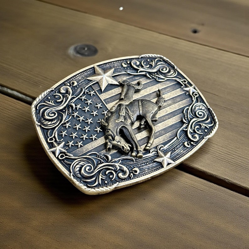 May include: A rectangular silver-toned belt buckle featuring a rodeo scene. The design includes a cowboy riding a bucking bronco against a backdrop of the American flag. The buckle is framed by decorative scrollwork and stars.