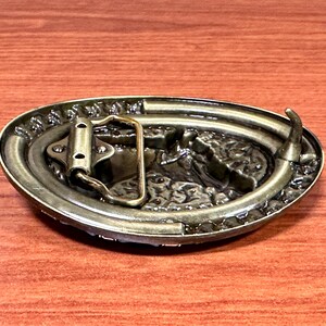 May include: An antique-style oval belt buckle with a detailed, raised design. The buckle is made of a bronze-coloured metal and features a textured border. The back of the buckle shows the belt loop and pin mechanism.