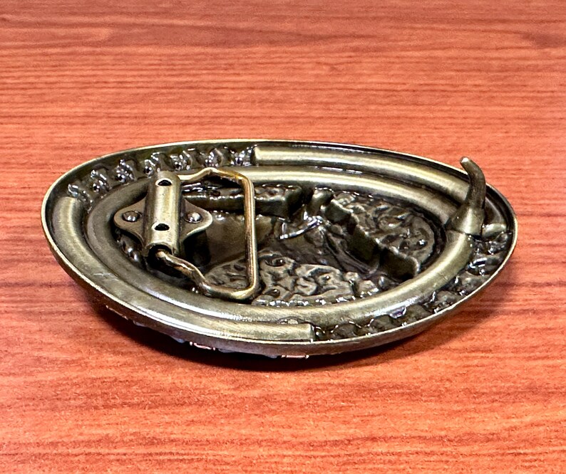 May include: An antique-style oval belt buckle with a detailed, raised design. The buckle is made of a bronze-coloured metal and features a textured border. The back of the buckle shows the belt loop and pin mechanism.