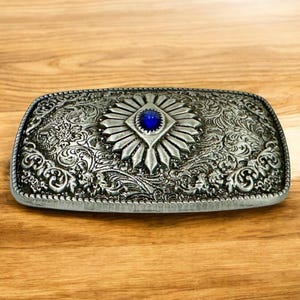 May include: Silver belt buckle with an ornate design and a blue gemstone in the center.