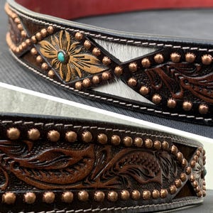 May include: Close-up of a handcrafted leather belt with intricate floral and geometric designs. The belt features copper studs, a turquoise accent, and a section of white and gray hair-on hide. The edges are stitched with white thread.