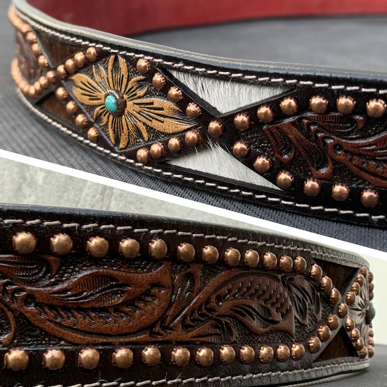 May include: Close-up of a handcrafted leather belt with intricate floral and geometric designs. The belt features copper studs, a turquoise accent, and a section of white and gray hair-on hide. The edges are stitched with white thread.