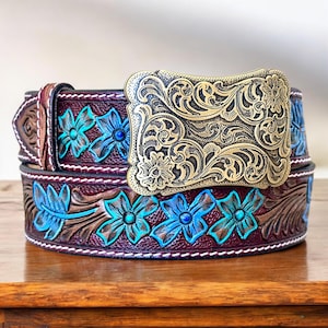 May include: A dark brown leather belt featuring a large, rectangular brass buckle with intricate floral scrollwork. The belt strap is embossed with a repeating pattern of flowers highlighted in bright turquoise paint and white stitching.