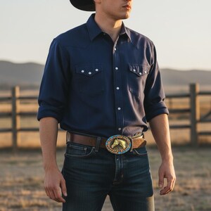 May include: A dark blue shirt with white buttons and a brown leather belt featuring a horse-themed buckle. The buckle has a gold horse head surrounded by colourful stones. The person is wearing blue jeans.