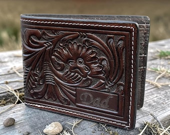 Puede incluir: Una cartera de cuero marrón oscuro con grabados florales y patrones en relieve. El panel frontal muestra la palabra Dad en una fuente cursiva, acentuada por costuras blancas en contraste a lo largo de los bordes del marco rectangular.