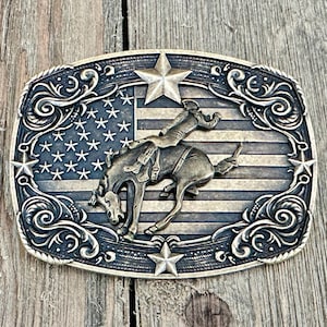 May include: A rectangular, antique-style belt buckle featuring a rodeo scene. The design includes a cowboy riding a bucking bronco against a backdrop of the American flag. The buckle is framed by decorative scrollwork and stars.