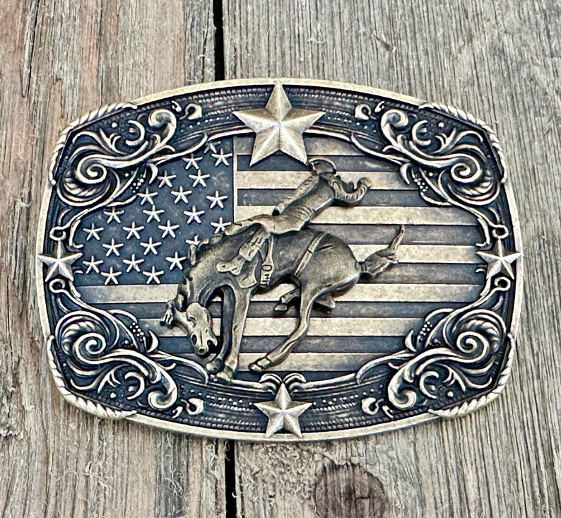 May include: A rectangular, antique-style belt buckle featuring a rodeo scene. The design includes a cowboy riding a bucking bronco against a backdrop of the American flag. The buckle is framed by decorative scrollwork and stars.