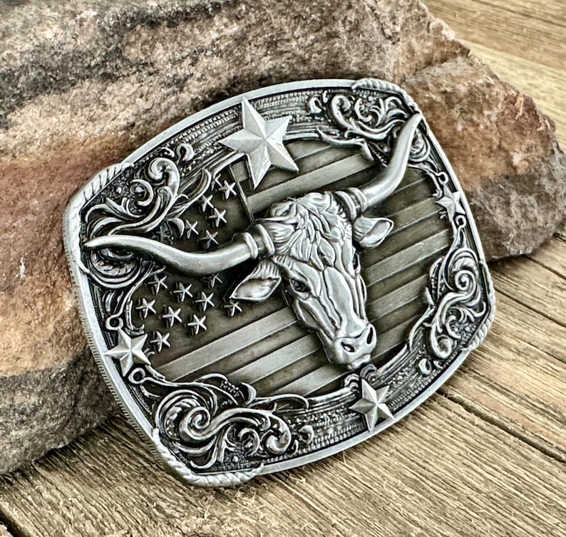 May include: Silver-toned metal belt buckle featuring a longhorn steer head with large horns, set against a backdrop of stars and stripes. The buckle has a rectangular shape with ornate scrollwork around the edges, and star accents.