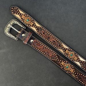 May include: Two brown leather belts with intricate floral and leaf tooling. One belt features a silver buckle and turquoise accents. The belts have a detailed design with copper-colored studs and a light-colored center.
