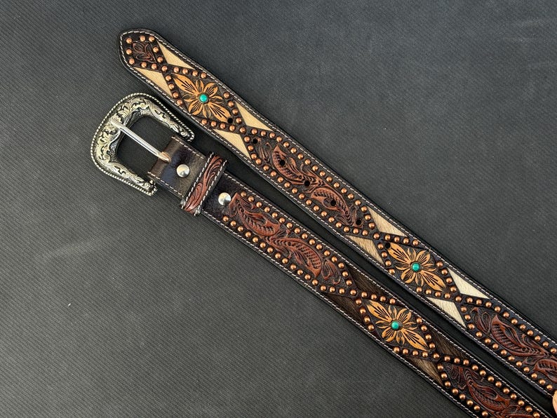 May include: Two brown leather belts with intricate floral and leaf tooling. One belt features a silver buckle and turquoise accents. The belts have a detailed design with copper-colored studs and a light-colored center.