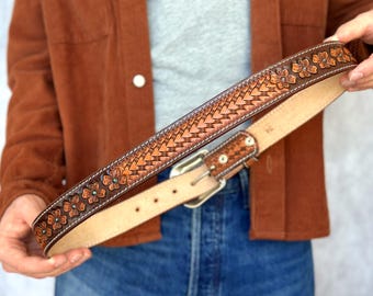 May include: A brown leather belt featuring a central embossed chevron pattern flanked by floral motifs with small turquoise-coloured centres. The belt has white contrast stitching along the edges, a silver-toned metal buckle, and a size 34 stamp.