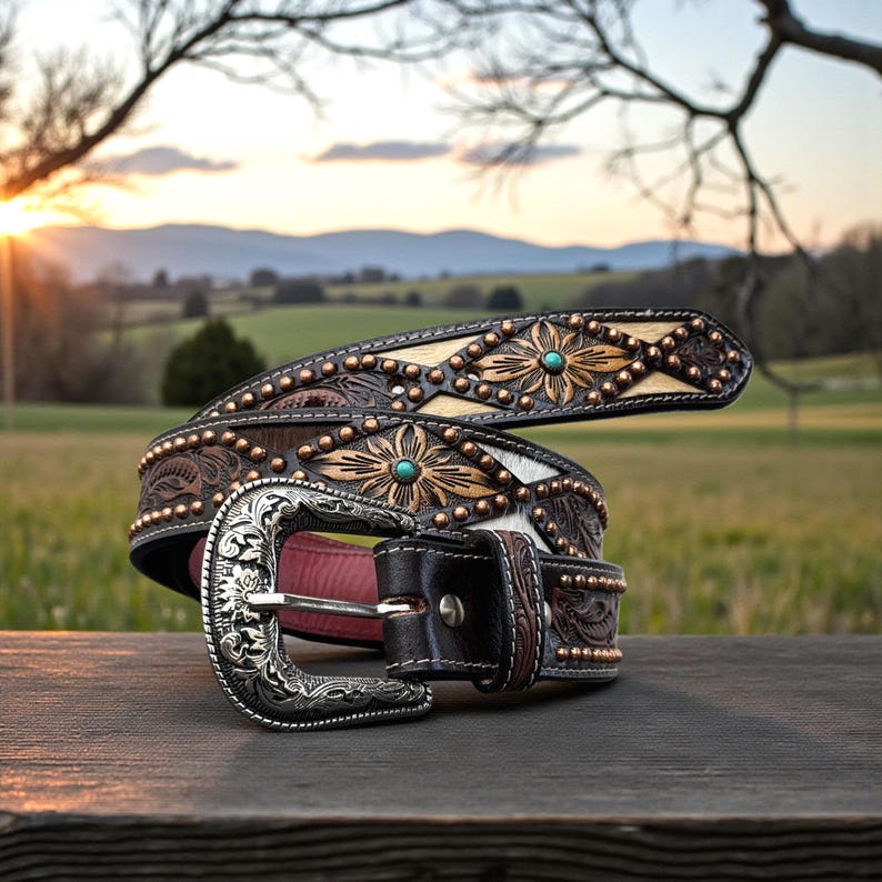 May include: A decorative brown leather belt with a silver buckle. The belt features floral and geometric patterns, turquoise accents, and copper studs. The background shows a sunset over a field and mountains.