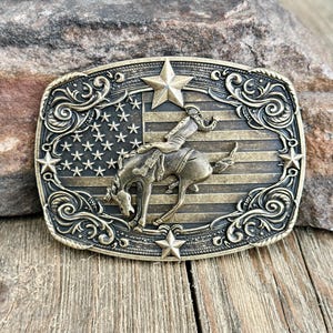 May include: A rectangular, antique brass belt buckle featuring a rodeo rider on a bucking bronco against a backdrop of the American flag. The buckle is adorned with stars and decorative scrollwork.