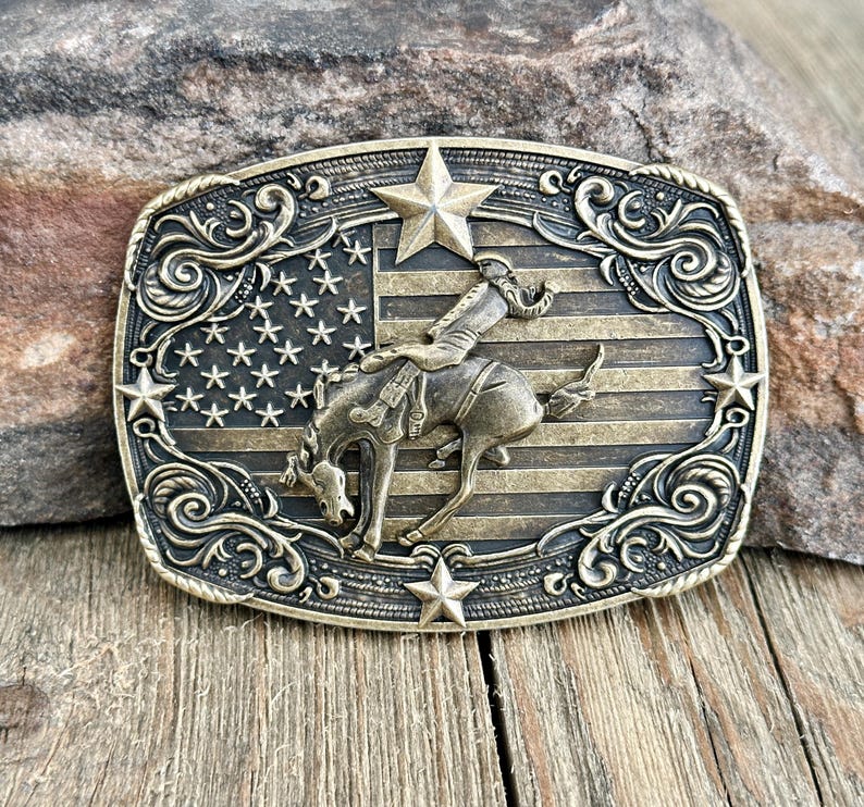 May include: A rectangular, antique brass belt buckle featuring a rodeo rider on a bucking bronco against a backdrop of the American flag. The buckle is adorned with stars and decorative scrollwork.
