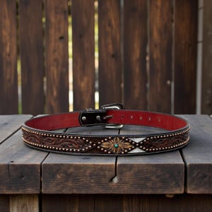 May include: A brown leather belt with a red interior. The belt features a decorative floral and leaf pattern with copper-colored studs and a turquoise center. The buckle is silver-colored.
