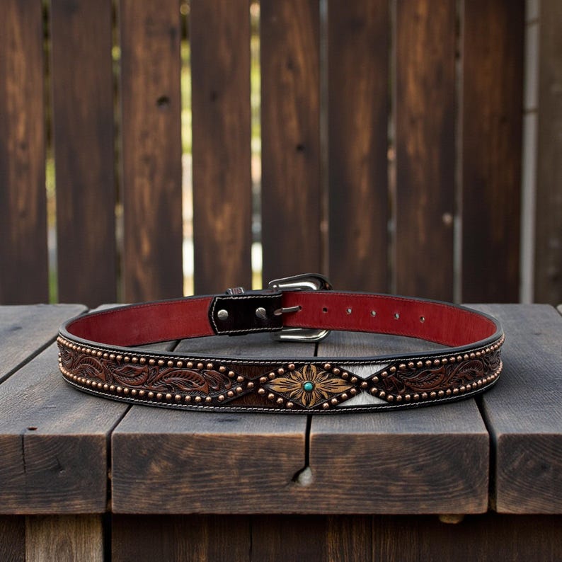 May include: A brown leather belt with a red interior. The belt features a decorative floral and leaf pattern with copper-colored studs and a turquoise center. The buckle is silver-colored.