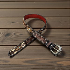 May include: A brown leather belt with intricate floral and leaf patterns, accented with small gold-colored studs and turquoise-colored stones. The belt has a silver-colored buckle and a red interior lining.
