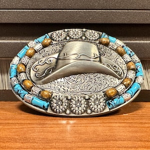 May include: Oval silver-tone belt buckle with a cowboy hat design. The buckle has turquoise and wooden bead accents around the border and floral details. The hat is in the centre of the buckle.